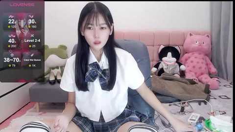 Media: Video of an Asian woman with long black hair, wearing a white short-sleeved shirt, plaid skirt, and knee-high striped socks, sitting on a pink chair in a messy room with stuffed toys and a TV screen displaying a game.