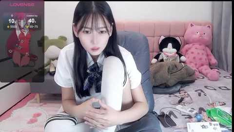 Media: A video of an Asian woman with long black hair, wearing a white shirt and plaid tie, sitting on a bed surrounded by stuffed animals and scattered items.