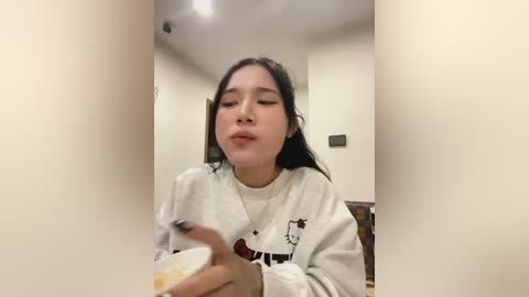 Media: Video of an Asian woman with long black hair, wearing a white sweatshirt, eating soup from a bowl in a dimly lit, beige-walled room.