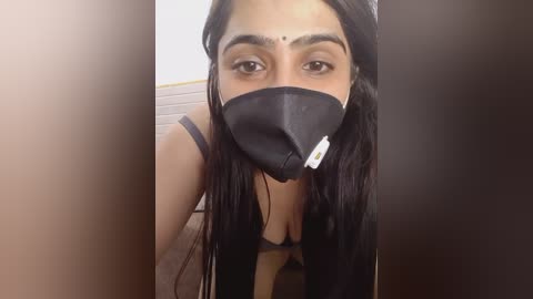 Media: Video of a young woman with medium-brown skin, long black hair, and a black face mask, wearing a black bra. She has dark eyebrows, a bindi, and is leaning forward, showing cleavage. Background shows beige tiles and a blurred wooden surface.