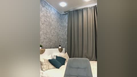 Media: Video of a modern, minimalist bedroom with a gray-patterned wallpapered wall, white headboard, teal pillows, a gray chair, and floor-to-ceiling gray curtains.