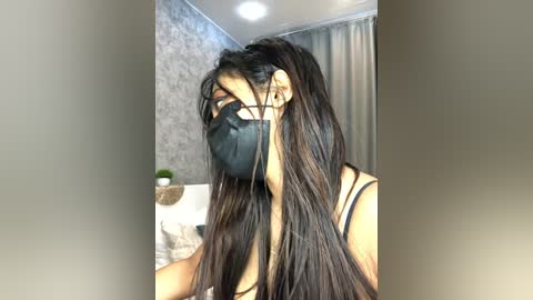 Media: Video of a young Asian woman with long, dark hair, wearing a black face mask, black tank top, and light makeup, sitting in a room with gray patterned wallpaper, beige curtains, and a white bed with pillows.