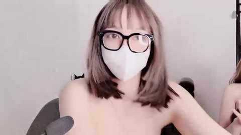 Media: Video of a light-skinned woman with shoulder-length brown hair, wearing black-rimmed glasses and a white face mask, sitting topless on a black chair. The background is plain and neutral.