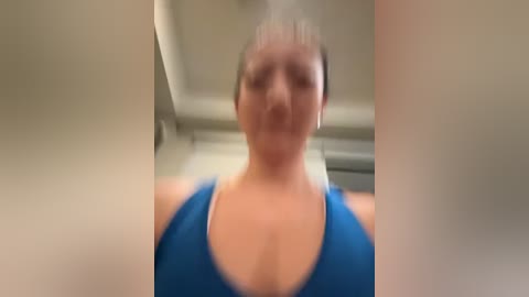 Media: A blurred video of a woman with a fair complexion, wearing a blue sports bra, standing in an indoor setting with beige walls and a ceiling fan.