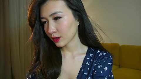 Media: Video of a young Asian woman with long, dark hair, fair skin, and red lipstick, wearing a navy blue dress with white floral patterns, seated indoors against a beige wall and mustard-yellow couch.