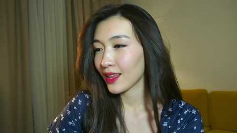 Media: Video of an East Asian woman with long black hair and fair skin, wearing a dark blue floral dress. She has a gentle smile and is in a softly lit room with beige curtains and a mustard-yellow couch in the background.