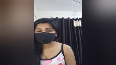 Media: Video of a woman with long black hair, wearing a black face mask and a sleeveless top with pink hearts, standing in a room with dark curtains and white walls.