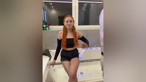 Media: Video of a young, fair-skinned woman with long, straight red hair, wearing a black off-shoulder crop top and matching shorts, standing in a modern, minimalist bathroom with a white tub and large windows.