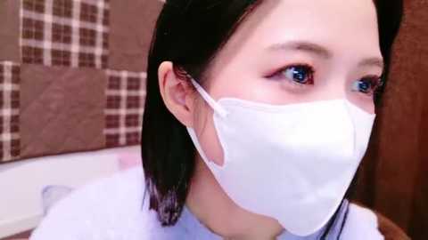 Media: Video of a young Asian woman with shoulder-length black hair, wearing a white surgical mask and a light blue top, in a bathroom with tiled walls.