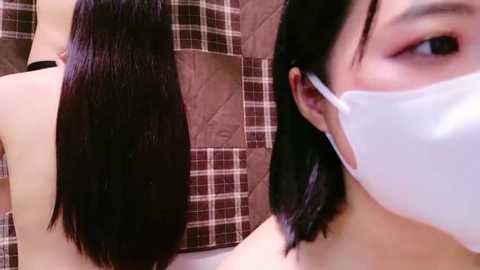 Media: A video of a woman with long, straight black hair and light skin, wearing a white surgical mask, against a plaid brown and white quilted background.