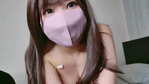 Media: Video of a young Asian woman with long, straight brown hair, wearing a lavender mask and no shirt, revealing her bare chest, in a minimalist bedroom with white walls and a black headboard.