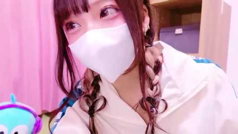 Media: Video of an Asian woman with brown hair in braids, wearing a white surgical mask, light blue hospital gown, and pink curtains in the background.