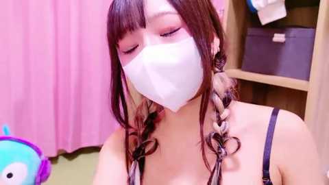 Media: Video of an Asian woman with long, brown hair styled in twin braids, wearing a white face mask, black spaghetti straps, and a pink backdrop.