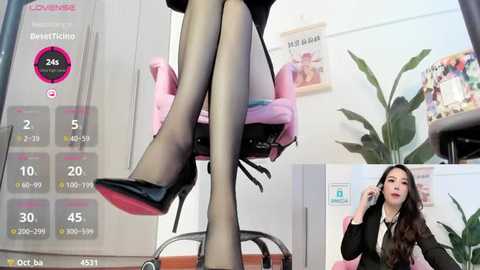 Media: Video of a woman in a black blazer, black tights, and black heels, sitting in a pink office chair. A virtual reality display in the background shows her legs, and another woman speaking on a phone.