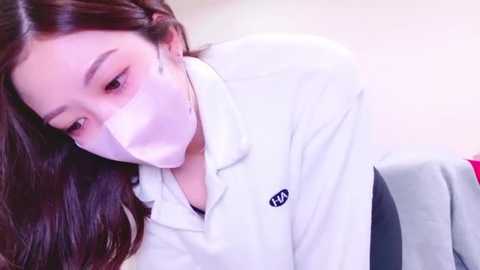 Media: A video of a young Asian woman with long black hair, wearing a white surgical mask and a white lab coat, leaning forward, possibly examining something, with a soft-focus background.