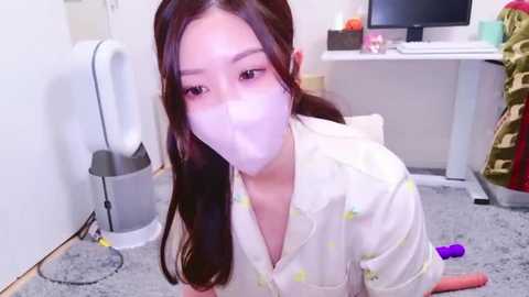 Media: Video of an Asian woman with long brown hair, wearing a white robe with yellow patterns, sitting on a grey carpet, face masked, in a cluttered office.