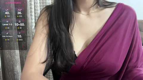 Media: Video of a light-skinned woman with long, straight black hair, wearing a deep purple one-shoulder dress with a black lace bra visible. Background features a patterned curtain.