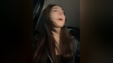 Media: A blurred video of a young woman with long brown hair, wearing a black jacket, sitting in a dimly lit car, mouth slightly open, possibly in mid-speech or mid-laugh.