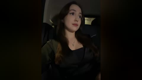 Media: Video of a young woman with long brown hair, wearing a black dress, seated in a dark, dimly-lit car interior, focused on a book, reading intently.