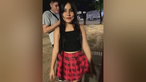 Media: A video shows a young woman with long dark hair, wearing a black crop top and red plaid skirt, smiling on a dimly lit street. A man in a grey shirt is behind her, partially visible.