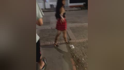 Media: Video of a young woman with long dark hair, wearing a black top and red skirt, walking on a wet pavement, reflected in a puddle, blurred background, dim lighting.