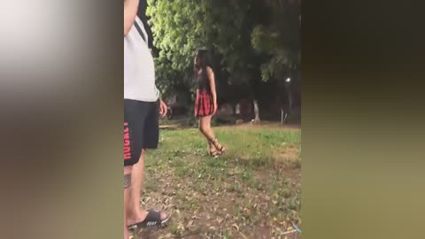 Media: Video of a woman in a red plaid skirt and black top chasing a man in a white tank top and black shorts on a grassy field with trees in the background.