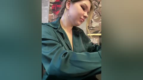 Media: Video of a young Caucasian woman with light skin, wearing a dark green leather jacket, sitting with arms crossed, looking downward. Background features abstract artwork with muted tones.