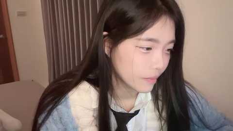 Media: A close-up video of a young East Asian woman with long black hair and a sad expression, wearing a light jacket over a striped shirt, indoors with vertical blinds in the background.