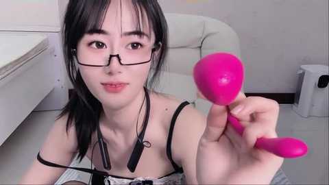 Media: A video of an Asian woman with straight black hair, wearing glasses, holding a bright pink dildo. She is in a minimalist, white room with a white chair and a white cabinet.