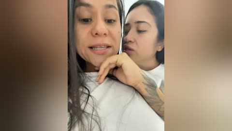 Media: Video of two young women with medium skin tones, one with long dark hair and a tattoo on her arm, the other with short black hair. They are wearing white tops, smiling, and appear to be in a casual, intimate setting.