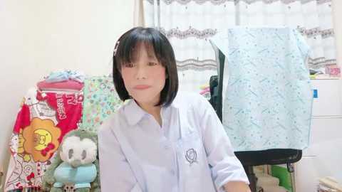 Media: A video of a young Asian woman with straight black hair and a pale complexion, wearing a white short-sleeve shirt. The background features a colorful, patterned wall with stuffed animals and a light blue blanket.