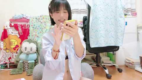 Media: A video of an East Asian woman with straight black hair, wearing a white shirt, eating a sandwich in a cluttered room with plush toys, a desk, and a chair.