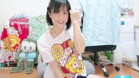 Media: Video of an East Asian woman with straight black hair, smiling while talking on a phone, wearing a white t-shirt with cartoon characters. Background features stuffed toys, a desk chair, and colorful fabric.