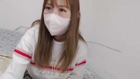 Media: Video of an Asian woman with long, straight, light brown hair, wearing a white long-sleeve shirt with red stripes, a white mask, and glasses, lying on a gray and white bedspread.