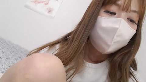 Media: Video of a young Asian woman with long, straight brown hair, wearing a white mask and white top, sitting on a white bed with speckled gray linens, and a framed baby photo on the white wall.