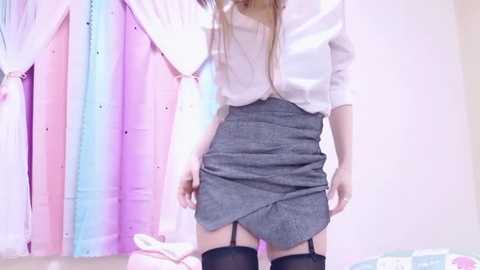 Media: Video of a slender woman in a white blouse, grey skirt, and black stockings, standing against a pastel, gradient-colored backdrop.