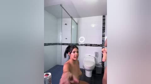 Media: Video of a nude woman with dark hair, small breasts, and light skin in a modern, minimalist bathroom with white walls and black mosaic tiles, standing in front of a white toilet.