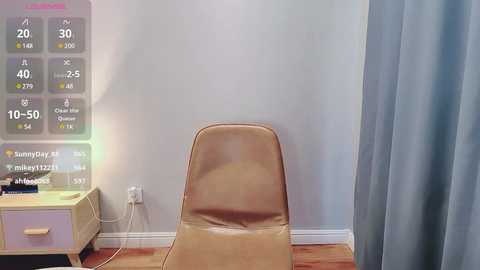 Media: Video of a modern room with a beige leather chair facing a light gray wall, a light wooden side table with a lamp, and a blue curtain partially drawn.