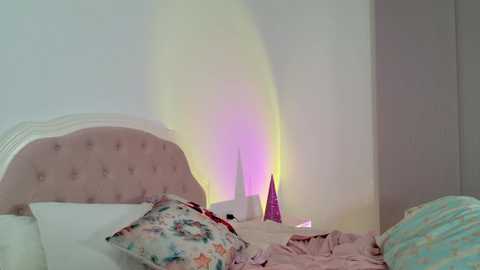 Media: A video of a serene bedroom with a white headboard, pastel-colored pillows, and a soft pink quilt. A colorful light display projects a vibrant yellow, pink, and blue glow on the wall, adding a whimsical touch.