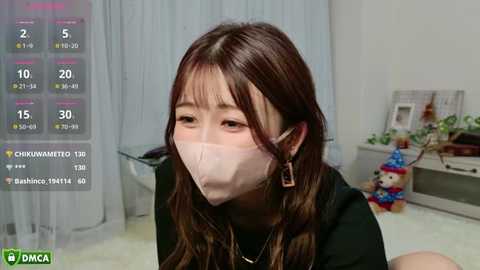 Media: A video of an East Asian woman with long brown hair and bangs, wearing a white mask, black top, and earrings, sitting indoors, surrounded by a virtual reality game interface.