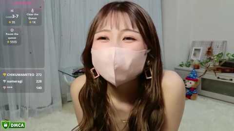 Media: Video of an Asian woman with long brown hair, wearing a pink face mask, smiling, in a brightly lit room with white walls, plants, and a calendar.