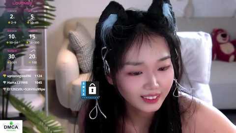 Media: Video of a young Asian woman with long black hair, wearing cat ears and a red top, smiling in a cozy room with a couch and plants.