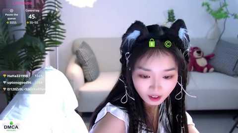 Media: Video of an Asian woman with long black hair, wearing cat ears, in a modern living room with a gray couch, plant, and stuffed toys.