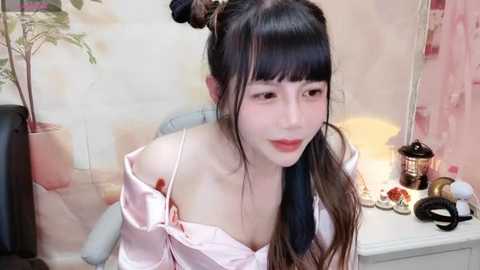 Media: Video of an Asian woman with pale skin and black hair in pigtails, wearing a pink satin robe, sitting at a desk with makeup, plants, and a laptop in the background.