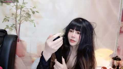 Media: A video of a young Asian woman with long black hair, fair skin, and a slender physique, wearing a black top, using a smartphone in a softly lit room with a potted plant and a lamp in the background.