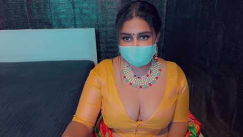 Media: Video of a South Asian woman with medium-dark skin, wearing a yellow blouse, gold necklace, and a turquoise mask, sitting on a dark couch against a dark brick wall.