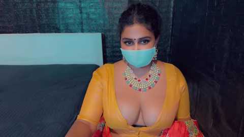 Media: A video of a South Asian woman in a mustard-yellow blouse, revealing cleavage, wearing a face mask, sitting on a bed with dark curtains in the background.