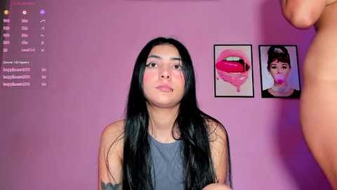 Media: Video of a Latina woman with long black hair, medium build, wearing a grey tank top, standing in a pink room with two framed lips paintings on the wall.