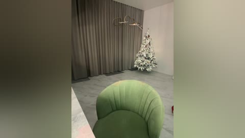 Media: A video of a minimalist room with a green, curved chair in the foreground, a white Christmas tree with snowflakes in the background, and grey curtains.