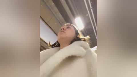 Media: A video of a young woman with light skin and brown hair, wearing a white, fluffy bathrobe, standing in a modern, industrial bathroom with metallic fixtures and a bright light overhead.